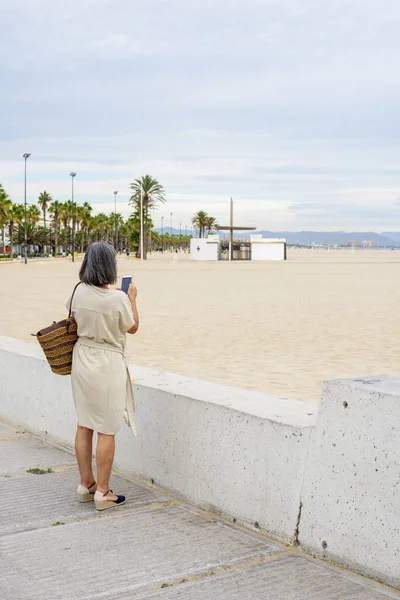 一个优雅的女士站在石墙旁边给大海拍照的背影 — 图库照片