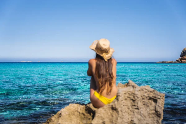 夏日的晴天 一个戴着帽子和太阳镜的小女孩坐在碧绿静谧的海滨岩石前 — 图库照片