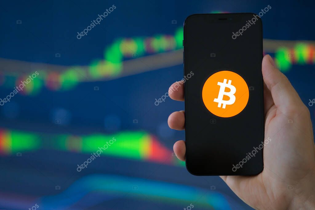 DUESSELDORF, GERMANY - Jul 10, 2021: Close up shot of Bitcoin logo on smartphone with stock candlesticks in the background