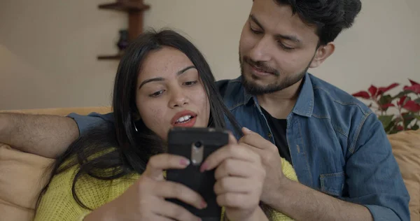 Young Indian Couple Happily Chatting While Sitting Sofa Using Phone — 스톡 사진
