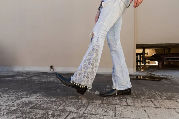 A closeup of a person wearing leather boots with heels and flare pants while walking on the pavement