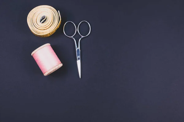 A flat lay of scissors, measuring tape, and thread on a dark background with free space for text