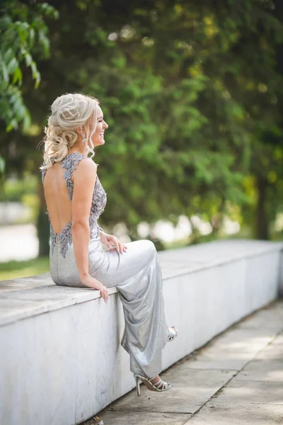 銀のドレスを着た幸せな若い女性が腰を開いてベンチに座って — ストック写真