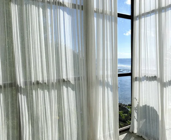 A beautiful view of beach house window with white tulle curtains with ocean view on a sunny day