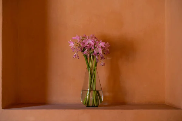 Wilting and dying pink or purple flowers in a glass vase, in an alcove on a ochre terracotta orange wall, symbolising sadness, loss, time, entropy.