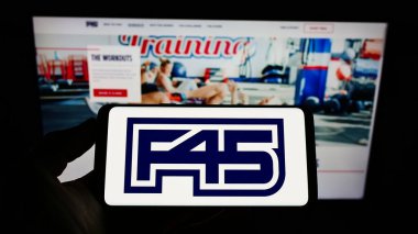 STUTTGART, GERMANY - Jul 12, 2021: Person holding cellphone with logo of fitness company F45 Training Holdings Inc. on screen in front of business webpage. Focus on phone display. clipart