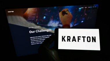 STUTTGART, GERMANY - Jun 14, 2021: Person holding mobile phone with logo of South Korean video games company Krafton Inc. on screen in front of web page. Focus on phone display. clipart