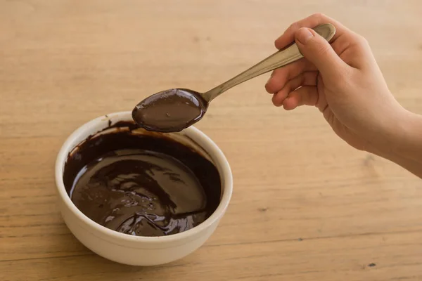 一只手从木制桌子上的白色陶瓷碗里拿着勺子融化的巧克力的镜头 — 图库照片