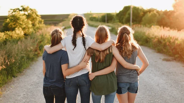 A back view of a group of female friends hugging and walking outdoors on a sunny day