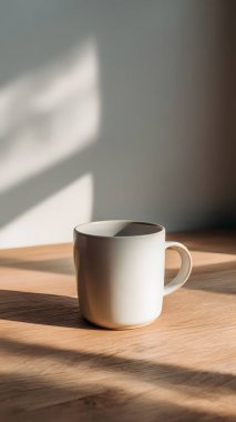 Temiz beyaz bir kupa, ahşap bir yüzeyde, yumuşak, rahatlatıcı gölgeler ve atmosfer yaratan, doğal güneş ışığıyla aydınlatılmış, zarif bir şekilde oturmaktadır..