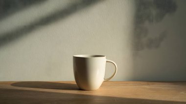Temiz beyaz bir kupa, ahşap bir yüzeyde, yumuşak, rahatlatıcı gölgeler ve atmosfer yaratan, doğal güneş ışığıyla aydınlatılmış, zarif bir şekilde oturmaktadır..