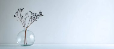 Zarif kurumuş çiçek sapları şeffaf, yuvarlak bir vazoda duruyor. Sakin, minimalist ve sanatsal bir oda atmosferi yaratıyor..