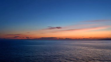 Uzun pozlama fotoğrafları, gün doğumunda ya da batımında çarpıcı bir şekilde renkli bir gökyüzünün altında, soluk, alçak bir kara ya da dağ silueti ile mavi, turuncu ve pembe bulutlardan oluşan yatay şeritler içeren engin ve sakin bir okyanus yüzeyi yakalar.