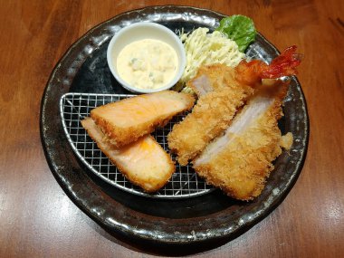 Kızarmış somon ve karides Japon yemeği, lahana salatası sebzesi ve sos sosu siyah tabakta servis edilir.