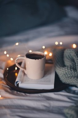 Bir fincan taze kahve, açık katlanmış kitabın üzerinde. Yatakta, ahşap tepside duruyor. Yakın çekim. İyi sabahlar. Kış tatili. Sıcak ev atmosferi. 