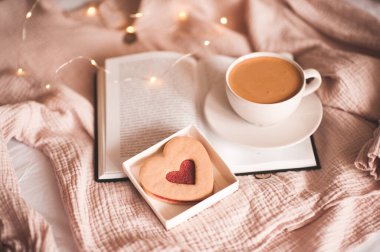 Bir fincan kahve, ışıkların üzerinde kalp şeklinde kurabiye olan açık bir kitabın üzerinde duruyor. İyi sabahlar. Kahvaltı vakti geldi. Sevgililer Günü.