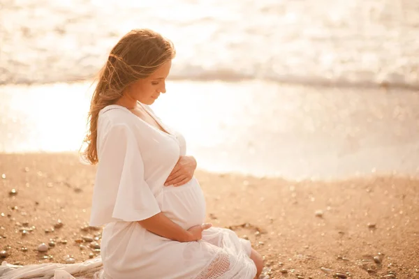 怀孕的女人穿着白色的衣服坐在海滨户外 健康的生活方式 图库图片