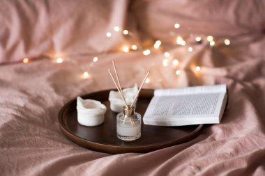 Sıvı ev parfümü cam şişede ve ahşap çubuklar tepside mumlar ve yatakta açık kağıt kitap. İyi sabahlar. Rahatlama. Seçici odak.