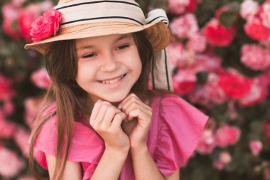 5-6 yaşlarında sevimli bir kız çocuğu açık havada bahçe gülleriyle poz veren şapkalar ve yazlık elbiseler giyiyor. Yaz sezonu. Çocukluk. Doğa arkaplanı üzerinde yeni yürümeye başlayan şık çocuk. 