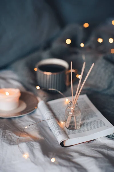 Bottle with liquid home fragrance and wooden bamboo sticks, scentes burn candle and cup of black tea over glow lights at background close up. Winter holiday season. Cozy romantic home atmosphere.