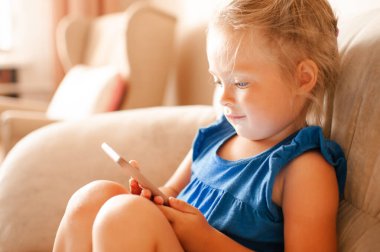 3-4 yaşlarında güzel bir kız çocuğu elinde akıllı bir telefonla oturma odasında çizgi film seyrediyor. Çocukluk. Teknoloji ile gülümseyen komik çocuk..