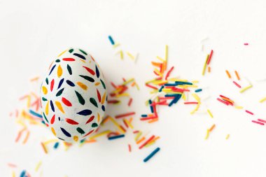 Bir Paskalya yumurtası kırmızı, sarı, mavi gouache ile beyaz zemin üzerine boyanmış, çok renkli şeker çubukları serpiştirilmiş. Yukarıdan bir fotoğraf. Bir yazıt için yer.