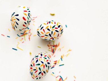 Üç Paskalya yumurtası beyaz zemin üzerinde kırmızı, sarı, mavi boya ile boyanır, odak noktasından çok renkli şeker çubukları serpiştirilir. Yukarıdan bir fotoğraf. Bir yazıt için yer.