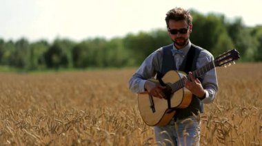 Güneş gözlüklü genç yakışıklı gitarist sarı bir alanda klasik gitar çalıyor. Spikeletler rüzgarda hareket eder. Gitarist parmaklarıyla telleri çalar. Arka planda, bulanık yeşil ağaçlar.