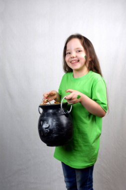 Aziz Patrick Günü tatili. Ellerinde ve yanağında yeşil yonca yaprağı olan yeşil elbiseli güzel, gülümseyen küçük kız leprikon döküm demir tenceresini gösteriyor. Altın dolu.