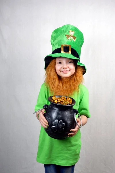 Saint Patricks Day holiday. Pretty smiling small girl in green clothes with decorative red beard and leprechaun hat, green shamrock leaf on her cheek, showing off cast iron lepreaun pot, full of gol