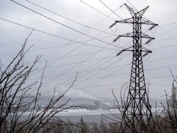 On a high mountain, a power line tower rises. Branches of trees, a river and the snow-covered opposite bank of the river with a populated area are visible.