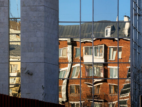 The mirrored surface of the building reflects an old red brick multi-storey building and part of a yellow building. The reflection of the building is distorted, refracted and looks unreal.