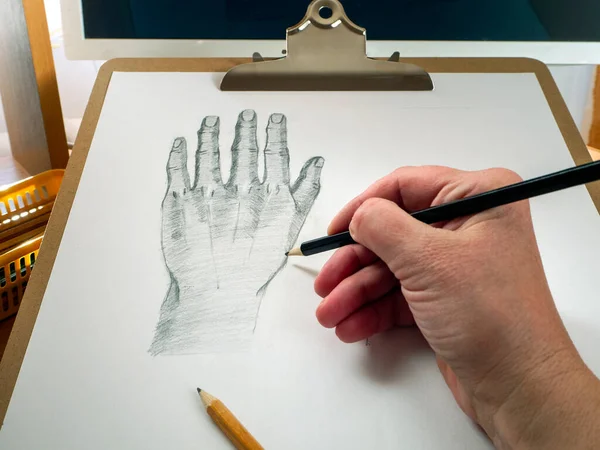 Pencil hand draws hand on paper. There is a monitor behind the drawing tablet. Concept of online education, online drawing lessons.