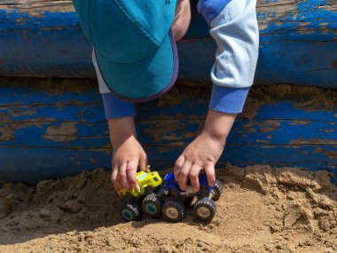Küçük bir çocuğun kulplarında birbirlerine doğru yönelmiş farklı renkte iki çocuk oyuncak arabası vardır. Çocuk boyalı kütüklere doğru eğildi ve arabaları kumda yuvarladı..
