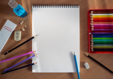 An open, blank sketchbook sheet is positioned vertically. A set of colored pencils, pencils separately, a sharpener and rubber bands are laid out side by side. Clean space for text.
