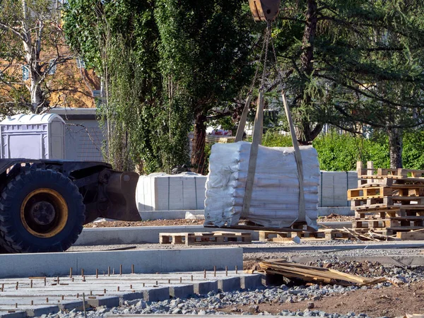 The crane transports the packed cargo. Nearby unfinished sidewalk, building materials, wooden coasters. Road construction works.