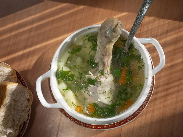 Chicken soup with vegetables, herbs and chicken drumstick is poured into a bowl. Pieces of bread are lying side by side on a plate. Background with homemade appetizing soup.