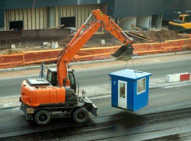 Tekerlekli bir kazıcı kabin taşır. Kazı makinesi inşaat alanından geçiyor. Arka planda bitmemiş bir evin duvarı var. Endüstri ve iş geçmişi.