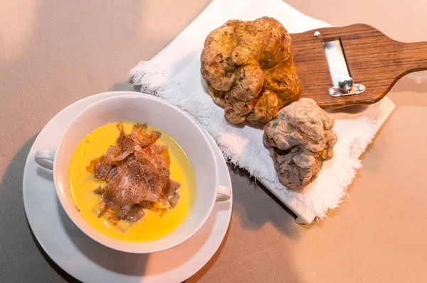 two big white alba truffles on wooden slicer and white napkin near cheese fondue with slices of truffle in white cup, in top view