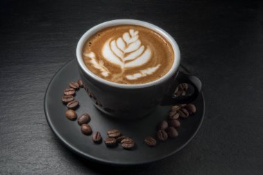 Siyah bardakta kahve, köpüklü yaprak resmi ve çay tabağında kahve çekirdekleri, siyah kayrak taşı arka plan.