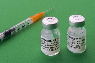 TURIN, ITALY - 27 Temmuz 2021: Pfizer-BioNTech COVID-19 Aşı Comirnaty şişeleri, yeşil arka planda şırıngası olan iki orijinal aşı şişesi