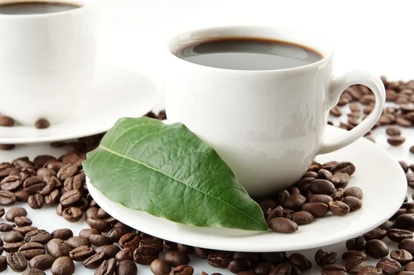 Scattered coffee beans with cups of coffee and leaf — Stock Photo, Image