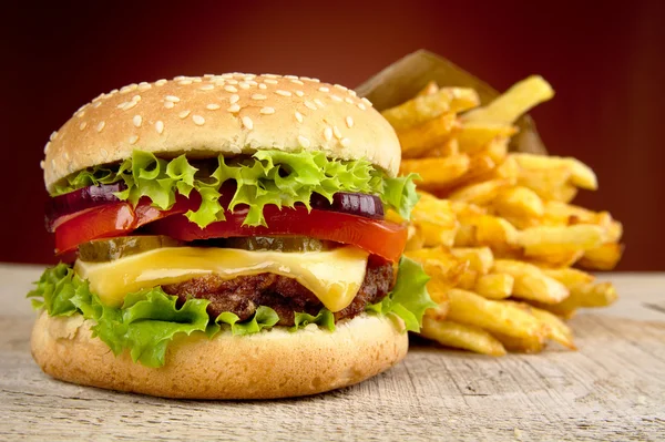 Big cheeseburger with french fries in bag paper on wooden table on red dpotlight background — Stock Photo, Image