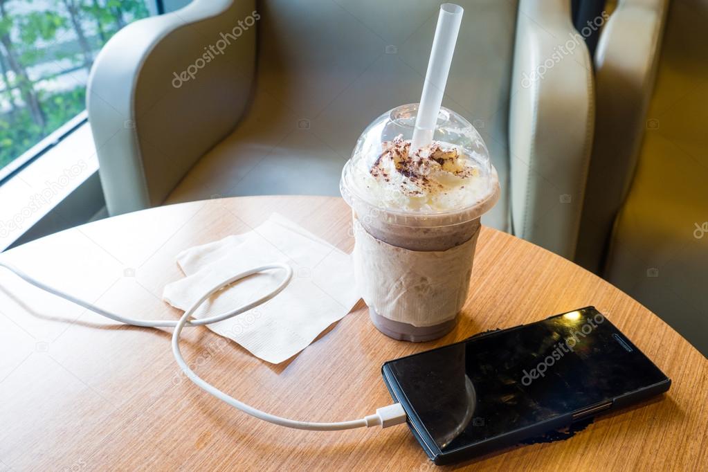 Cell phone charging in the cafe with a plastic cup of iced chocolate frappe