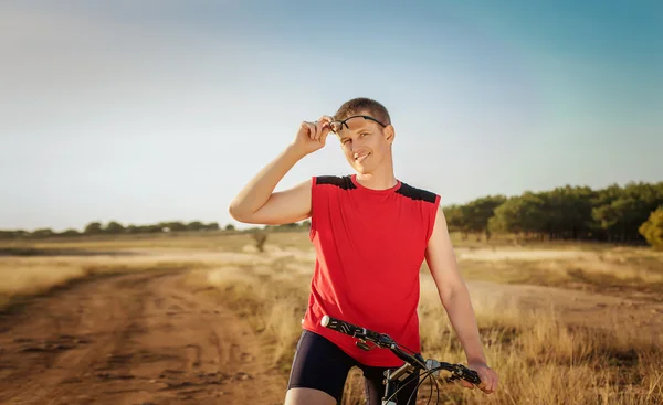 Joven hombre guapo montando una bicicleta en el campo sobre un fondo de cielo azul — Foto de Stock