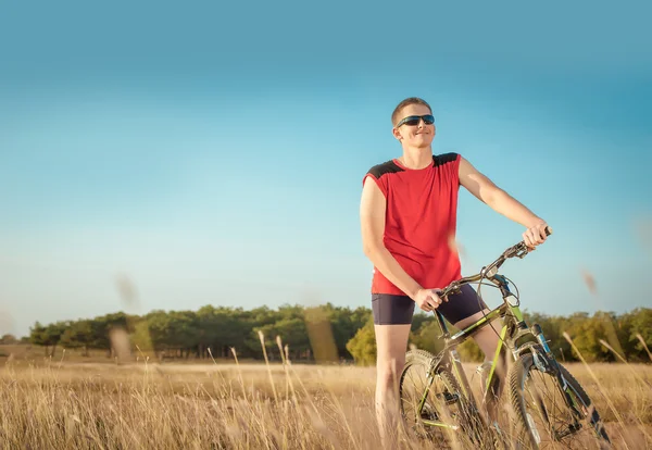 Young handsome man riding a bicycle in the field on a background of blue sky — Stok fotoğraf