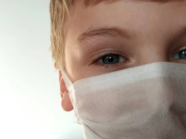 Masked child. The face of a 7-year-old boy in a protective white surgical mask close-up. Schoolboy with blond hair and blue-gray eyes on white background.