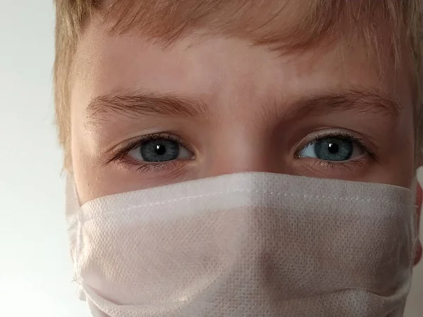 Masked child. The face of a 7-year-old boy in a protective white surgical mask close-up. Schoolboy with blond hair and blue-gray eyes on white background.