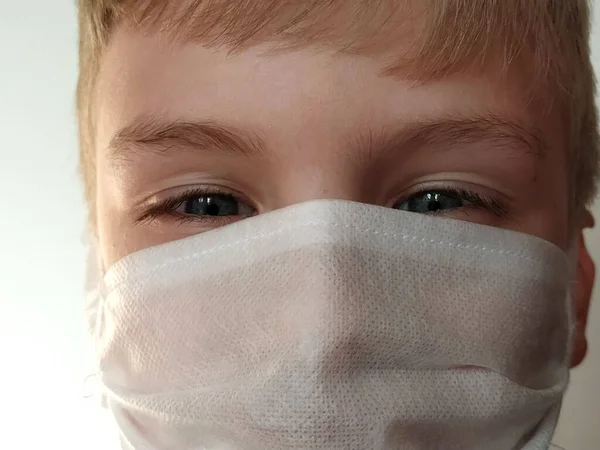 Masked child. The face of a 7-year-old boy in a protective white surgical mask close-up. Schoolboy with blond hair and blue-gray eyes on white background.