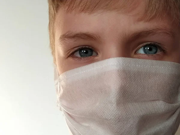 Masked child. The face of a 7-year-old boy in a protective white surgical mask close-up. Schoolboy with blond hair and blue-gray eyes on white background.